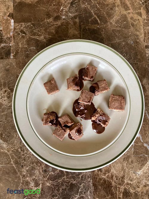 Small bite-sized chocolate brownies arranged on a white plate with a marble tabletop. Some brownie pieces have melted chocolate on them.